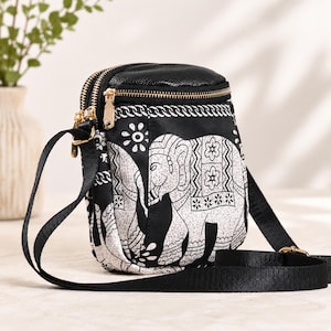 May include: A small black crossbody bag featuring a white elephant print with floral and geometric patterns. The bag has a textured black top panel, two gold-tone metal zippers, and an adjustable black fabric shoulder strap.