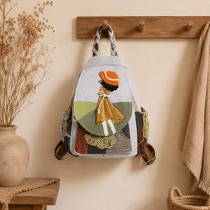 May include: A light gray backpack featuring a braided top handle and a decorative applique of a figure wearing an orange hat and a mustard yellow dress. The lower section displays a dark fabric with a fan-shaped leaf pattern in gold and gray.