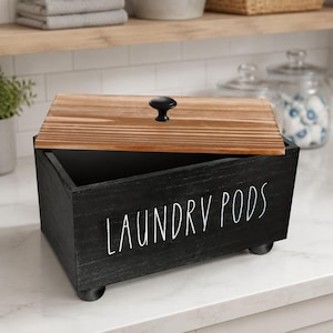 Può includere: Una scatola rettangolare in legno nero con la scritta LAUNDRY PODS in bianco. La scatola presenta un coperchio in legno naturale con un piccolo pomello nero e poggia su quattro piccoli piedini neri. Il design è semplice e funzionale.