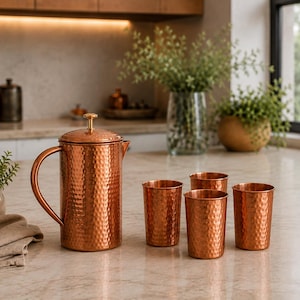 May include: A hammered copper pitcher with a matching lid and handle, accompanied by four matching hammered copper tumblers. The set features a distinct textured surface throughout, reflecting light across the metallic, reddish-brown finish.