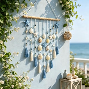 Pode incluir: Uma decoração de parede com uma haste de madeira horizontal e vários cordões brancos verticais. Os cordões são decorados com estrelas-do-mar azuis e brancas, várias conchas, pequenas contas azuis e brancas e borlas azuis e brancas.
