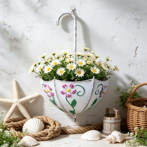 Puede incluir: Una maceta de pared de metal blanco con forma de paraguas invertido, con diseños florales en relieve de color rosa y verde. La maceta está llena de un denso arreglo de margaritas blancas con centros amarillos para decorar la pared.