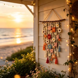 Pode incluir: Uma decoração de parede composta por um ramo de madeira horizontal com várias trepadeiras verticais de crochê verde, adornadas com pequenas flores de crochê em tons de branco, rosa, laranja, vermelho e azul, com borlas pendentes.