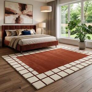 May include: A rectangular area rug featuring a solid rust-colored center panel surrounded by a cream-colored border with a grid pattern of thin rust-colored lines. The rug is placed on a wooden floor in a bedroom with a rust-colored upholstered bed.