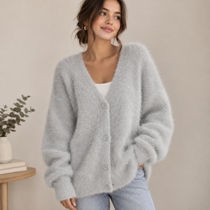 Puede incluir: Un cárdigan gris claro de textura esponjosa con cuello en V, mangas largas y cierre frontal de botones. La prenda de punto presenta una superficie suave y puños acanalados, combinada con una camiseta blanca y pantalones vaqueros.