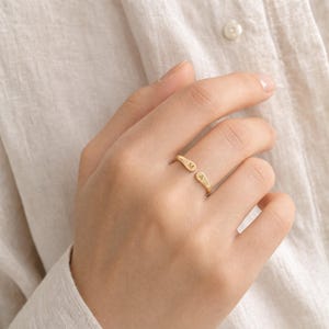 May include: A gold-toned open ring featuring two rounded ends, each engraved with a single uppercase letter. One end displays the letter M, while the other end displays the letter A. The ring has a polished, smooth finish and a simple, minimalist band.