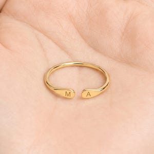 May include: A gold-toned open ring featuring a smooth, polished finish. The two ends of the band are slightly flattened and rounded, with the letter M engraved on one side and the letter A engraved on the other side of the opening.