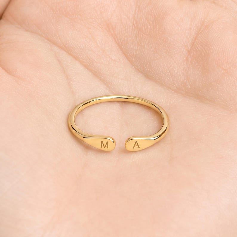 May include: A gold-toned open ring featuring a smooth, polished finish. The two ends of the band are slightly flattened and rounded, with the letter M engraved on one side and the letter A engraved on the other side of the opening.