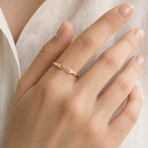 May include: A gold-toned open ring featuring two rounded ends, each engraved with a single letter. One end displays the letter M, while the other end displays the letter A. The band has a smooth, polished finish and a simple, minimalist design.