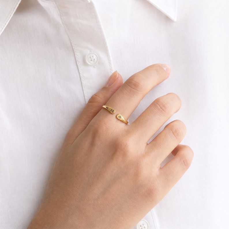May include: A gold-toned open ring featuring two tapered ends, each engraved with a single uppercase letter. One end displays the letter M and the other end displays the letter A. The ring has a polished metallic finish and a simple, slender band.
