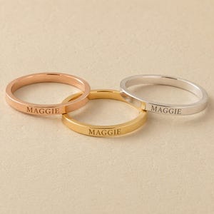 Peut inclure: Trois bagues fines en métal aux tons or rose, or jaune et argent. Chaque bague présente un design à anneau plat avec le nom MAGGIE gravé dans une police serif majuscule claire sur la surface extérieure. Les bagues sont disposées en groupe.