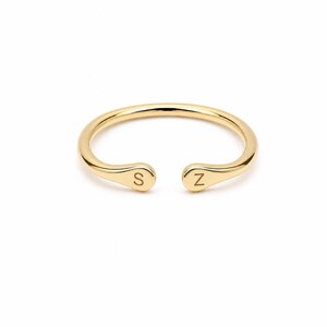 May include: A polished gold open ring featuring a smooth, rounded band. The two ends of the ring are slightly flattened and rounded, each engraved with a single capital letter. One end displays the letter S, and the other end displays the letter Z.