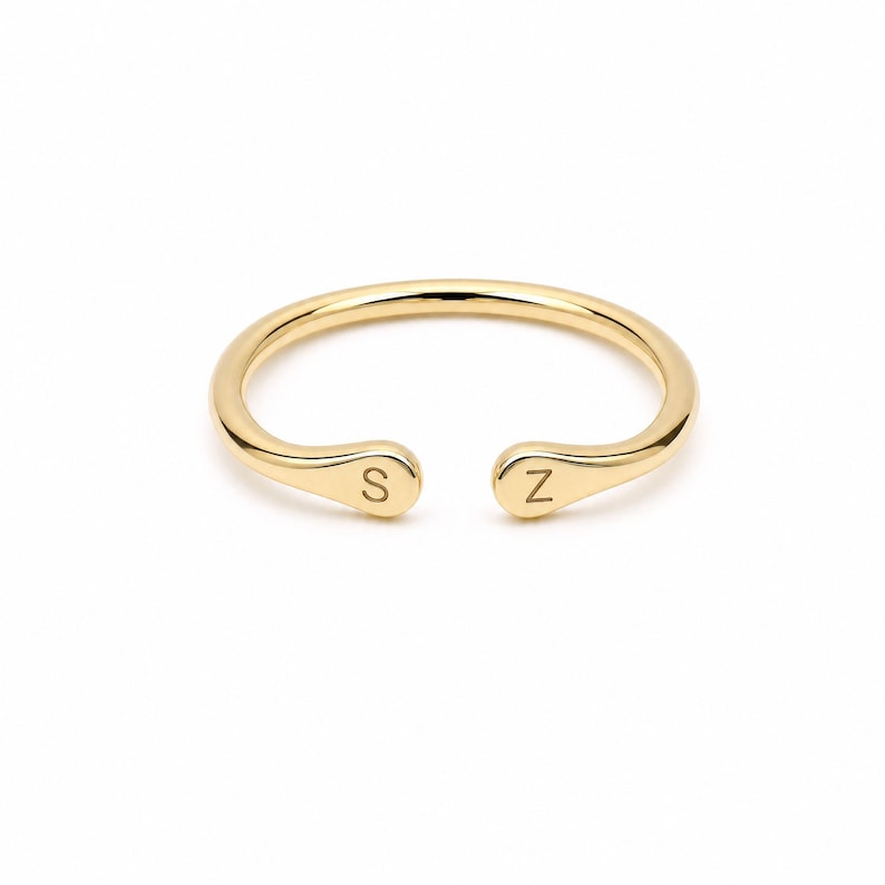 May include: A polished gold open ring featuring a smooth, rounded band. The two ends of the ring are slightly flattened and rounded, each engraved with a single capital letter. One end displays the letter S, and the other end displays the letter Z.