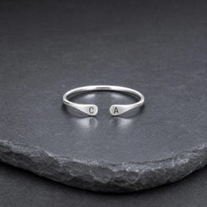 May include: A silver-toned open cuff ring featuring a smooth, polished finish. The two ends of the ring are flattened into small, rounded tabs, each engraved with a single uppercase letter, with the letter C on one side and the letter A on the other.