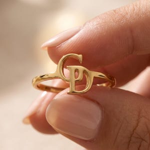 Op de afbeelding: Een goudkleurige ring met een gepolijste metalen band en een opvallend, gestileerd monogram als middelpunt, bestaande uit de in elkaar grijpende hoofdletters G en D, wat zorgt voor een doorlopend, glad en reflecterend oppervlak.