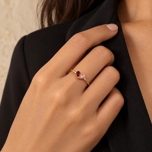 May include: A gold ring featuring two pear-shaped gemstones set side-by-side. One stone is a deep red colour, while the other is a soft, pale pink. The band is thin and polished, creating a delicate look that highlights the contrasting colours of the stones.