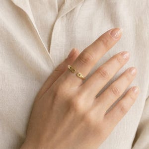 May include: A gold-toned open ring featuring two flat, rounded ends. Each end is engraved with a single uppercase letter, with the letter M on one side and the letter A on the other. The ring has a polished, smooth finish and a simple, adjustable band.