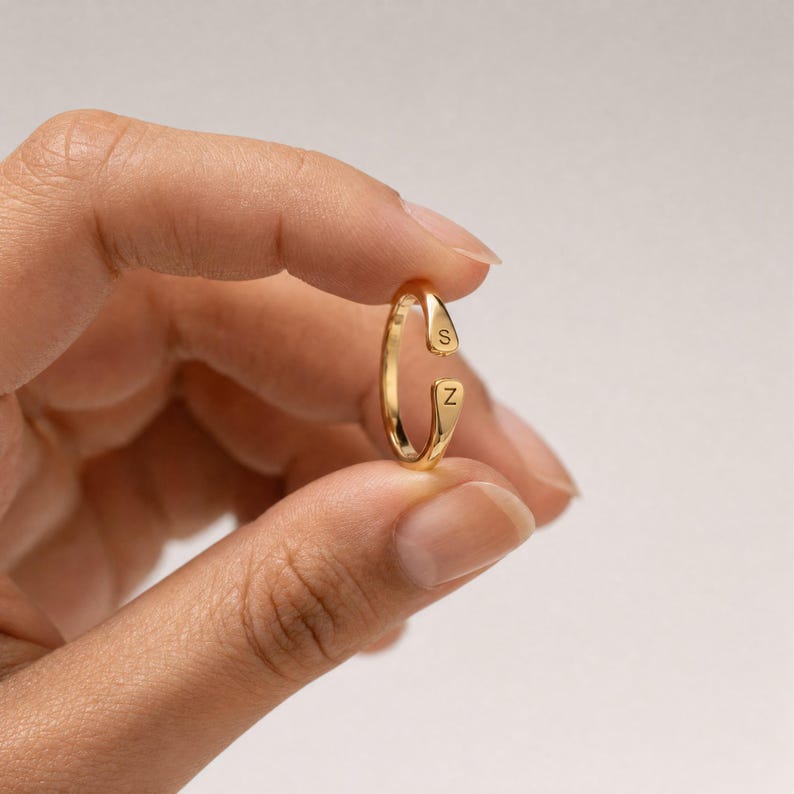 May include: A gold-toned open ring featuring two flat, tapered ends. Each end is engraved with a single uppercase letter, specifically the letter S on the top terminal and the letter Z on the bottom terminal, creating a personalised initial design.