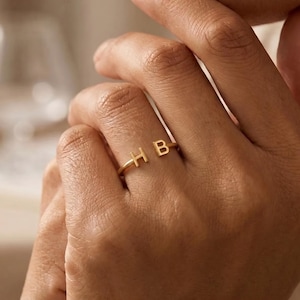 Op de afbeelding: Een goudkleurige open ring met de hoofdletters H en B aan de uiteinden van de band. De ring heeft een gladde, gepolijste afwerking en een dunne, eenvoudige metalen band die om de vinger sluit met een open ruimte tussen de twee letters.