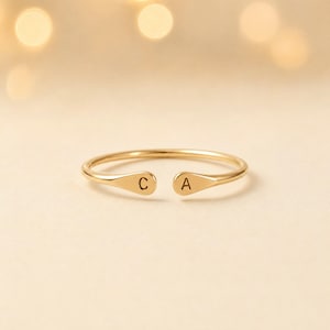Può includere: Un anello aperto color oro con due estremità piatte e arrotondate. Ogni estremità è incisa con una lettera maiuscola, con la lettera C sul lato sinistro e la lettera A sul lato destro. L'anello è sottile e presenta una finitura liscia e lucida.