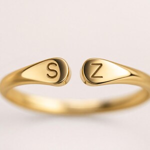 May include: A gold-toned open ring featuring two teardrop-shaped ends, each engraved with a single letter. The left end displays the letter S, and the right end displays the letter Z. The band has a smooth, polished finish throughout its structure.