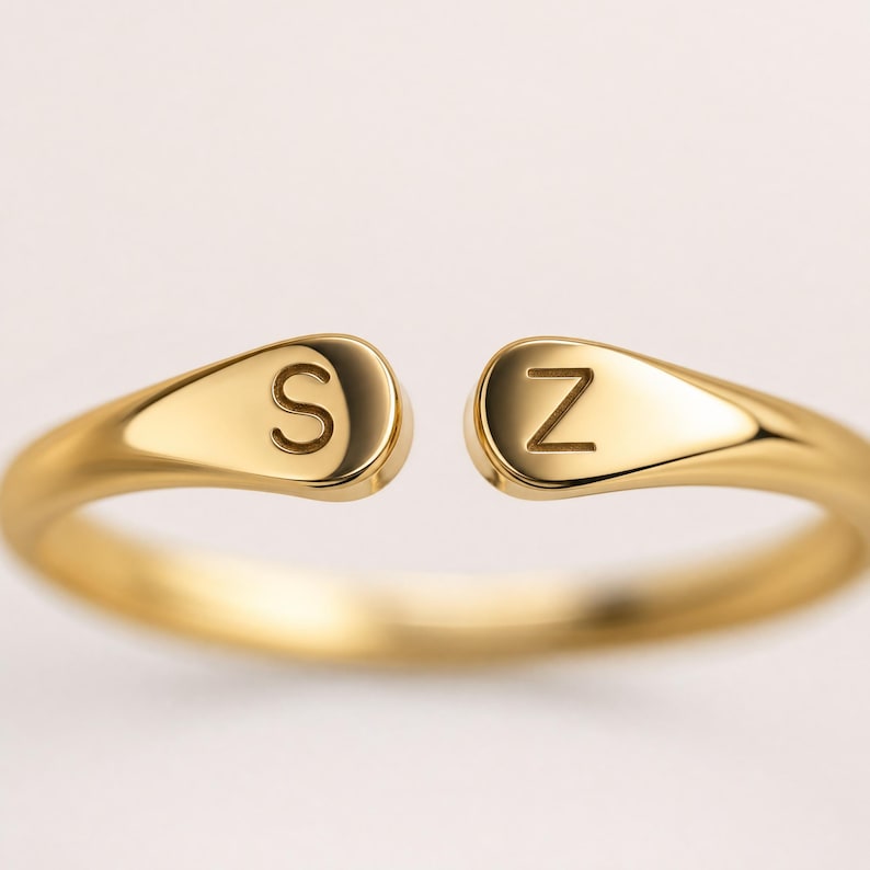 May include: A gold-toned open ring featuring two teardrop-shaped ends, each engraved with a single letter. The left end displays the letter S, and the right end displays the letter Z. The band has a smooth, polished finish throughout its structure.