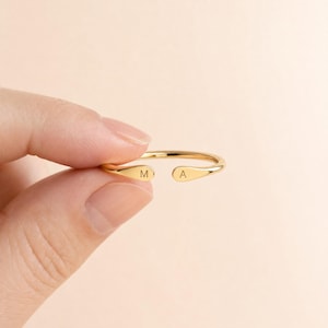 Può includere: Un anello aperto color oro con una finitura liscia e lucida, caratterizzato da estremità arrotondate e affusolate. L'estremità sinistra è incisa con la lettera M e quella destra con la lettera A, per un design personalizzato ed essenziale.