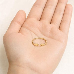 May include: A gold-coloured open ring featuring a thin, polished band. The two ends of the ring are slightly flattened and rounded, with the letter M engraved on one terminal and the letter A engraved on the other terminal, creating a personalised look.