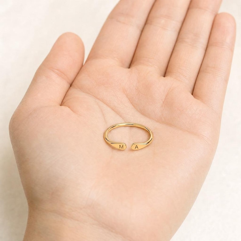 May include: A gold-coloured open ring featuring a thin, polished band. The two ends of the ring are slightly flattened and rounded, with the letter M engraved on one terminal and the letter A engraved on the other terminal, creating a personalised look.
