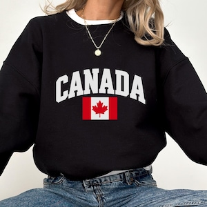May include: A black long-sleeve crewneck sweatshirt featuring the word CANADA in white arched capital letters above a graphic of the Canadian flag with a red maple leaf. The sweatshirt is worn over a white shirt and paired with blue denim jeans.