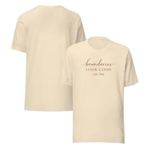 May include: A cream-colored short-sleeve t-shirt featuring the brown text "boundaries LOOK GOOD on me" printed on the front. The shirt is displayed from both the front and back angles, showcasing the plain back and the graphic design on the chest.
