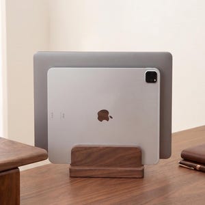 Könnte beinhalten: Ein rechteckiger, dunkelbrauner Holzständer, der ein silbernes Tablet und einen grauen Laptop in aufrechter Position hält. Der Ständer besteht aus glattem Massivholz und verfügt über einen horizontalen Schlitz für elektronische Geräte.