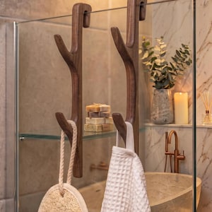 May include: Two dark brown wooden over-the-glass hooks with a branched design. Each hook features two prongs for hanging items. A round textured loofah with a rope loop hangs from one hook, and a white waffle-weave towel hangs from the other.