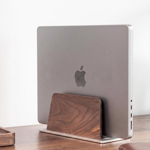 May include: A silver laptop with an Apple logo is held in a vertical position by a dark brown wooden stand. The stand features a smooth, natural wood grain finish and a rectangular base that supports the laptop in an upright, space-saving orientation.