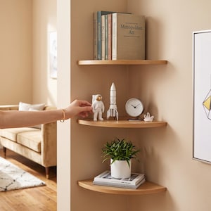 May include: Three light-toned wooden corner shelves mounted on a wall. The top shelf holds several books including one titled Metropolitan Art. The middle shelf displays an astronaut figurine, a rocket model, a clock, and a crystal. The bottom shelf holds a potted green plant and stacked books.