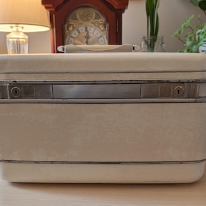 May include: A rectangular off-white hard-shell vanity case featuring a silver-toned metal trim, a top handle, and two integrated key locks on the front panel. The exterior has a smooth, solid finish with visible metallic accents along the center seam.
