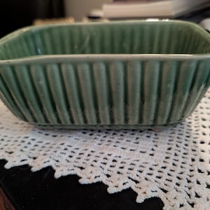 May include: A square, light green ceramic dish with vertical ribbed detailing. The dish has a slightly flared rim and sits on a white, open-weave doily. The dish is likely for serving or storage.