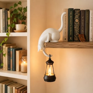 Peut inclure: Une figurine de chat blanc perchée sur le bord d'une étagère en bois, avec sa queue s'étendant vers le bas pour tenir une petite lanterne noire. La lanterne présente un boîtier en verre transparent et émet une lumière chaude et brillante.