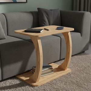 May include: A light wood C-shaped side table with a rectangular top and a matching base. The table features a curved support leg and a lower shelf holding a stack of three books. A black notebook and a pen rest on the table surface.