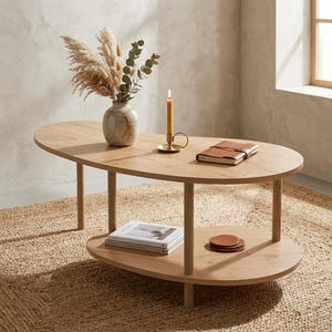 May include: A light wood coffee table with an oval top and a lower shelf. On the table sits a beige ceramic vase filled with dried pampas grass and eucalyptus, a gold candle holder with a yellow taper candle, and a brown leather-bound notebook.