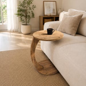 May include: A C-shaped side table crafted from light brown wood with a circular top and base, featuring a curved support column. A black mug sits on the tabletop surface, which is positioned over the edge of a beige upholstered sofa.