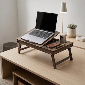 Puede incluir: Un soporte para portátil de madera marrón oscuro con una plataforma inclinable ajustable, una sección lateral plana con una ranura vertical para un smartphone, una hendidura rectangular para un teléfono y una pequeña ranura para un bolígrafo, con cuatro patas plegables.