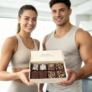 Op de afbeelding: Een rechthoekige kartonnen doos met acht vierkante pure chocolade brownies in twee rijen. De brownies zijn bestrooid met diverse zaden en kokosvlokken. Op het deksel staat de tekst "Willco Willow" en "No Added Sugar Protein Squares."