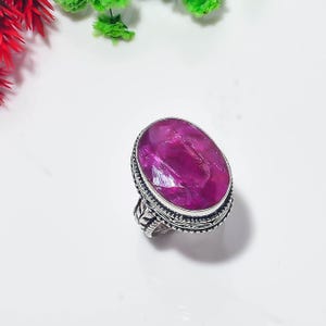 Puede incluir: Un anillo de plata con una gran piedra preciosa magenta de forma ovalada y tallada, engastada en un bisel de metal decorativo. La banda del anillo muestra un intrincado patrón de hojas en relieve en los laterales y detalles de cuentas.