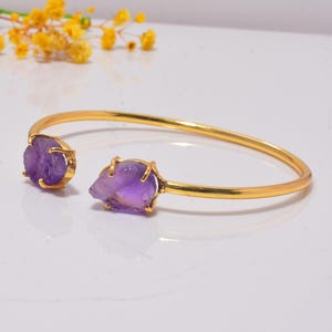 Könnte beinhalten: Ein goldfarbenes Armband in offener Form mit zwei rohen, violetten Amethyststeinen, die an beiden Enden in Krappenfassungen sitzen. Das Armband besteht aus einem glatten, polierten Metallband, das sich zu einem offenen Kreis biegt und die Textur betont.