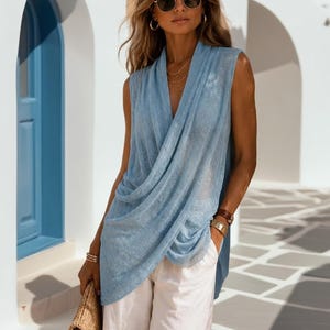 May include: A sleeveless light blue knit top with a draped cowl neckline and an asymmetrical hemline, paired with loose-fitting white trousers and a woven straw clutch bag. The top features a fine, textured knit fabric with a subtle sheen.