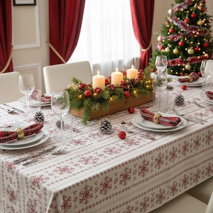 May include: A white tablecloth with a red snowflake pattern set for a festive meal. The table features a wooden centrepiece with three cream pillar candles, pine branches, red ornaments, and fairy lights, alongside plaid napkins with gold rings.