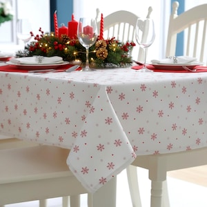 Op de afbeelding: Een wit tafelkleed met een herhalend patroon van kleine rode sneeuwvlokken en stippen. De stof ligt over een tafel die is gedekt met rode placemats, doorzichtige wijnglazen, witte borden en een middelpunt van dennentakken en rode bessen.