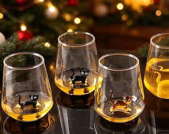 May include: Four clear glass tumblers, each containing a small, three-dimensional cat figurine at the bottom. Two glasses hold black cat figures, one holds a brown cat figure, and one holds a yellow cat figure, all filled with amber liquid.