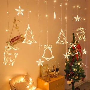 Peut inclure: Un ensemble de guirlandes lumineuses LED suspendues représentant des étoiles, des sapins de Noël, des cloches et des rennes. Une pancarte avec le texte Merry Christmas est accrochée au mur, près d'un petit sapin de Noël décoré.